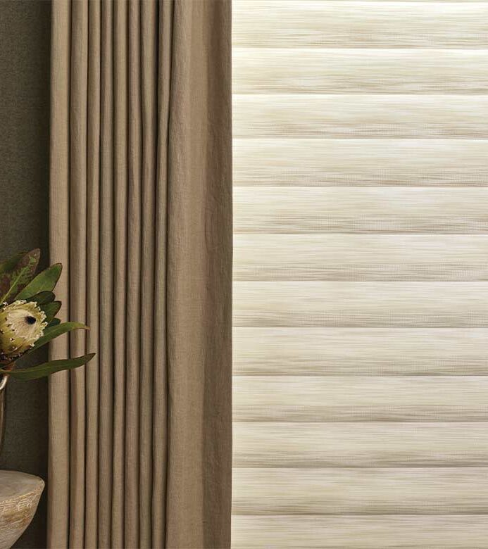 aura-illuminated-shades-detail-with-side-drapery-panels-hunter-douglas-bazaar-home-decorating close up of roman shade with drapery, enhanced with aura illuminated shades soft LED lighting